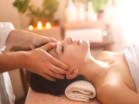 Spa therapist giving a woman a gentle facial massage in a calm, candlelit treatment room.