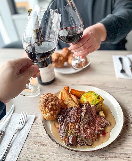 Guests toasting with red wine at a table set with a steak meal and sides.